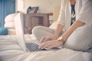 Woman sitting on a bed, using a laptop in a well-lit bedroom with a casual setting.