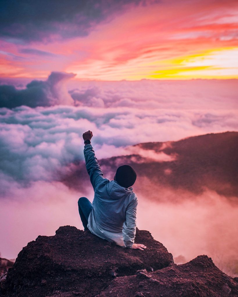 Person triumphantly raising fist atop a mountain at sunrise above clouds.