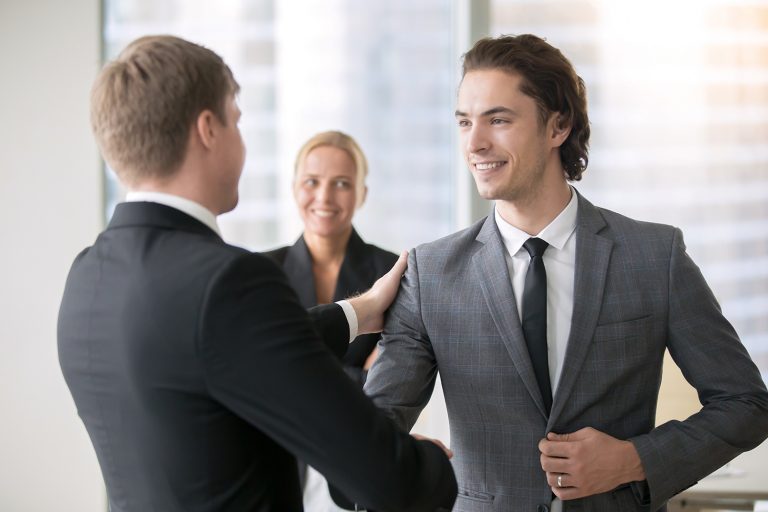 Two businessmen in suits shaking hands with a woman smiling in the background.