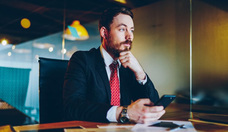 Thoughtful businessman in a suit holding a phone while sitting at a desk.