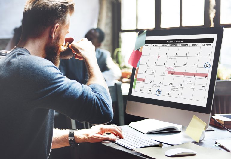 Man drinking coffee and working on a computer with a calendar displayed on the screen.