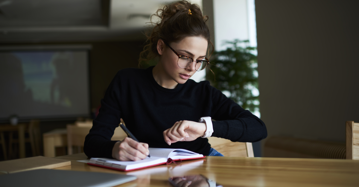 Focused woman checking watch while writing in notebook at desk.