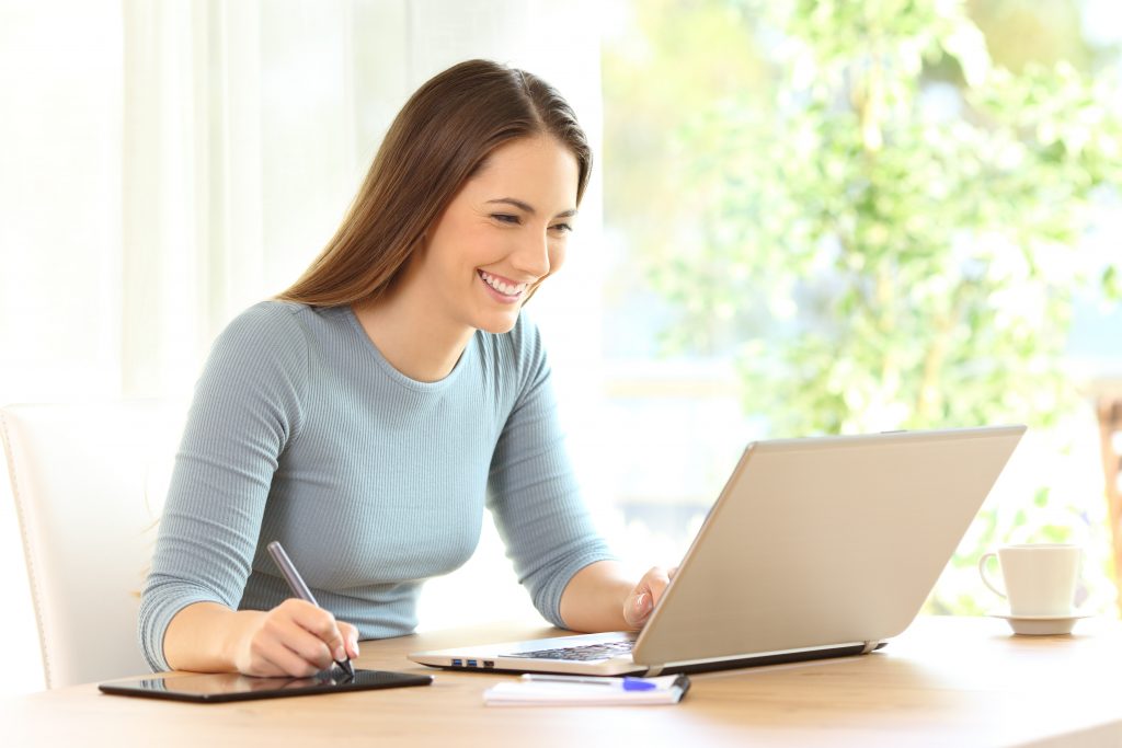 Smiling woman using laptop while writing notes, with coffee cup.