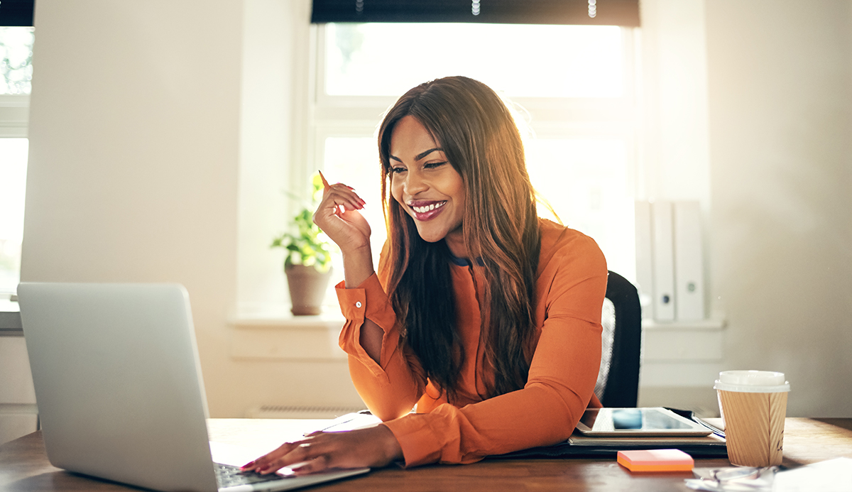Smiling woman in orange blouse working on laptop in a bright home office.