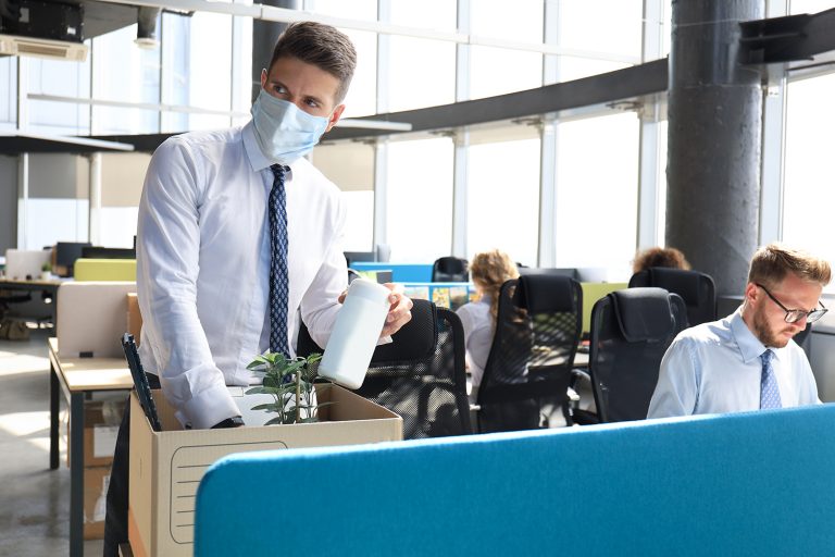 Man in a mask packing his belongings in a box at his office desk, with colleagues working in the background.