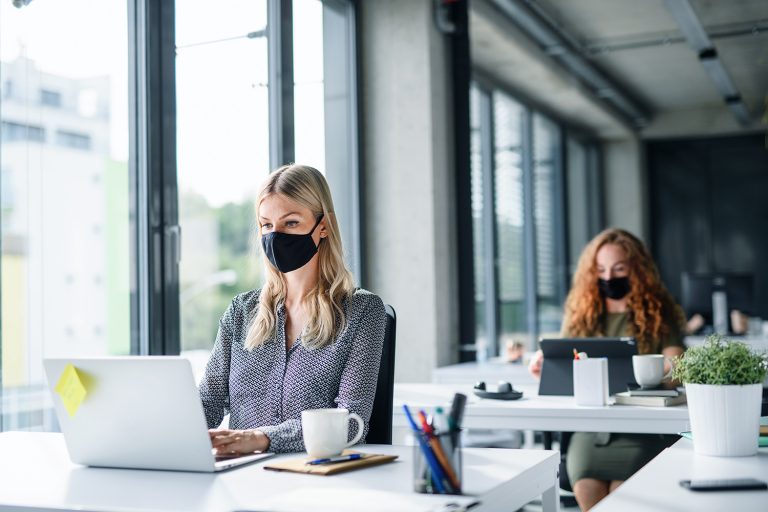 Two women wearing masks working diligently on laptops in a bright, modern office environment.