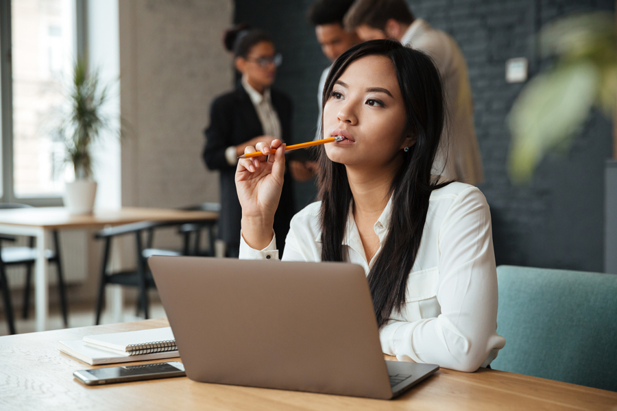 Professional Asian woman deep in thought while working on a laptop in a modern office setting.