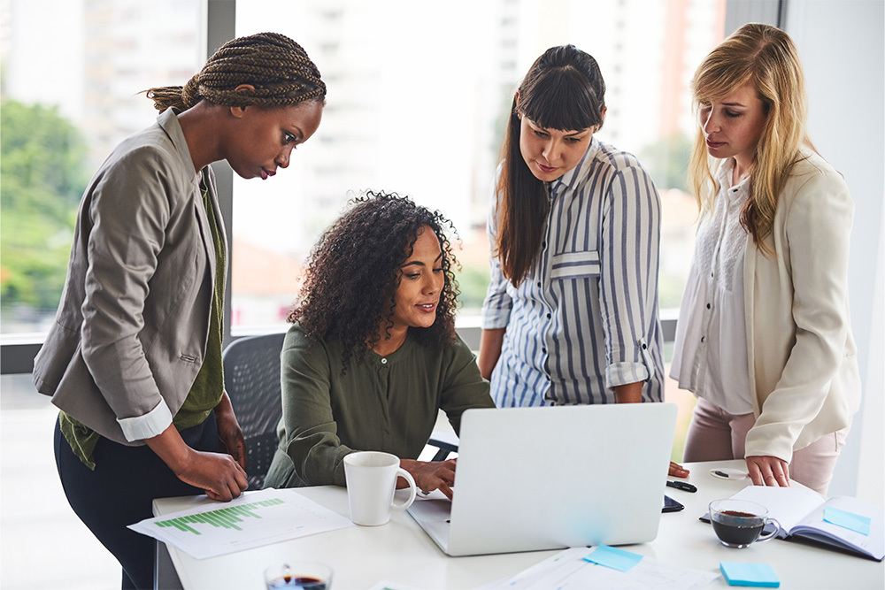 Group of professional women collaborating around a laptop in an office setting, showcasing teamwork and workplace collaboration.