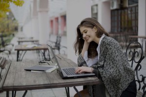 Happy young woman working on a laptop at an outdoor cafe table, smiling while reading the screen.